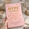 Pink journal titled 'The Hype Journal' held by a hand on a desk with decor items.